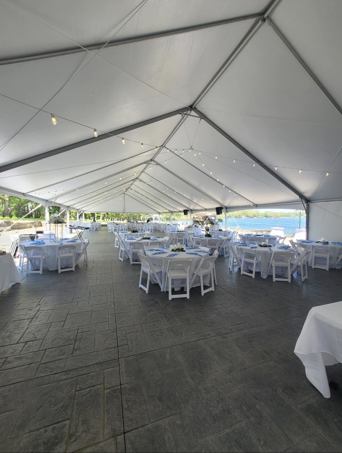 Elegant tented reception by the water with white linens, blue napkins, and string lights
